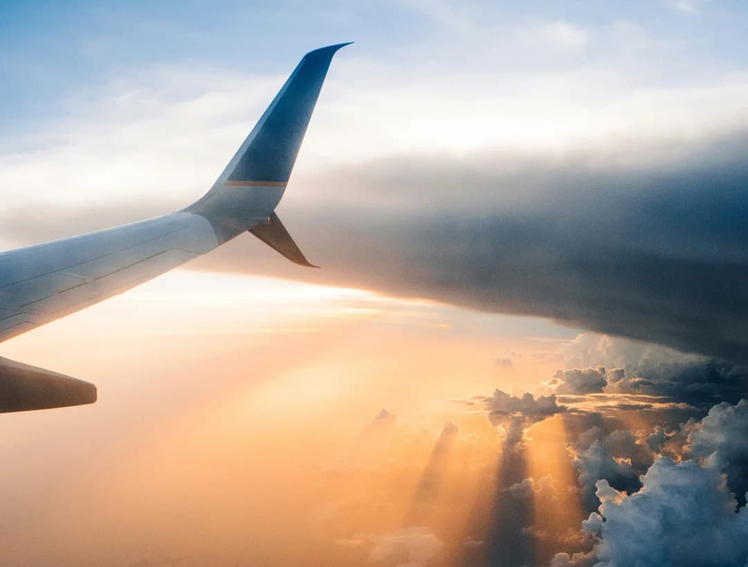 View from an airplane window showing the aircraft wing above a dramatic sunset sky, with golden light rays breaking through large storm clouds below.